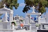 A cemetery with white marble tombstones and crosses, adorned with colorful flower arrangements. Statues, including one of Jesus, are visible amid tall trees. The blue sky and sea create a serene backdrop.