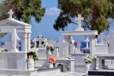 A cemetery with white marble tombstones and crosses, adorned with colorful flower arrangements. Statues, including one of Jesus, are visible amid tall trees. The blue sky and sea create a serene backdrop.
