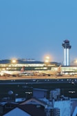 A bustling airport scene during evening hours with a prominent control tower. The terminal is brightly illuminated, revealing the architectural design and windows. Several large airplanes are parked on the tarmac, and airport vehicles are visible. The sky is a deep blue, indicating a clear atmosphere.