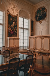 An opulent interior featuring ornate, gold-accented architectural details and classic artwork. A large, elegant table with mesh-backed chairs surrounds the center space. Large windows with sheer curtains diffuse natural light into the room. Decorative moldings and classical paintings adorn the walls.
