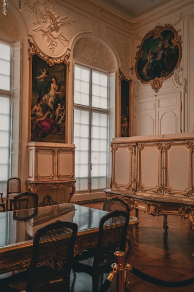 An opulent interior featuring ornate, gold-accented architectural details and classic artwork. A large, elegant table with mesh-backed chairs surrounds the center space. Large windows with sheer curtains diffuse natural light into the room. Decorative moldings and classical paintings adorn the walls.