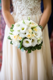 A person holding a bouquet of flowers with white and yellow blossoms, surrounded by lush green leaves. The floral arrangement is intricate and delicate, with a focus on the soft textures and natural elements of the blooms. The outfit worn by the person includes a richly detailed fabric with floral patterns.