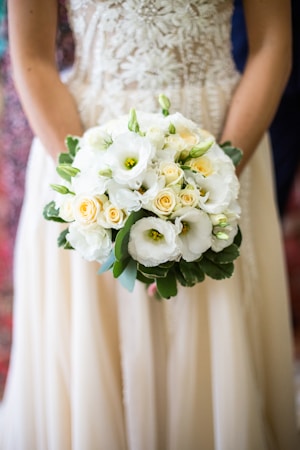 A person holding a bouquet of flowers with white and yellow blossoms, surrounded by lush green leaves. The floral arrangement is intricate and delicate, with a focus on the soft textures and natural elements of the blooms. The outfit worn by the person includes a richly detailed fabric with floral patterns.