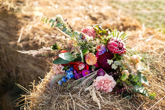 A vibrant bouquet of flowers rests on a bed of straw outdoors, featuring a mix of colorful blooms including pink peonies, red dahlias, and blue anemones, surrounded by green foliage and decorative grasses. The natural setting adds a rustic charm.