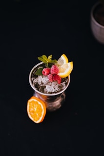 A refreshing cocktail garnished with a sprig of mint, two lemon slices, and three raspberries sitting atop crushed ice. The drink is presented in a clear glass with an additional slice of orange placed on the table beside it. The background is dark, highlighting the vibrant colors of the drink and its garnish.