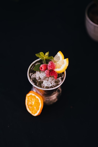 A refreshing cocktail garnished with a sprig of mint, two lemon slices, and three raspberries sitting atop crushed ice. The drink is presented in a clear glass with an additional slice of orange placed on the table beside it. The background is dark, highlighting the vibrant colors of the drink and its garnish.