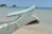 A close-up of a wooden boat prow with weathered paint in light blue, featuring some rust and peeling areas. The background shows a sandy beach and clear, calm ocean water with a few small islands and boats in the distance under a clear blue sky.