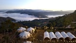 A serene and picturesque campsite is nestled among rolling hills and lush greenery. White tents are set up on a flat area surrounded by tall trees and decorative string lights, with a breathtaking view of a sprawling valley below. The valley is filled with a sea of clouds, and distant mountain peaks are visible against the horizon at sunrise or sunset, adding to the tranquility of the scene.
