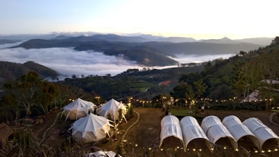 A serene and picturesque campsite is nestled among rolling hills and lush greenery. White tents are set up on a flat area surrounded by tall trees and decorative string lights, with a breathtaking view of a sprawling valley below. The valley is filled with a sea of clouds, and distant mountain peaks are visible against the horizon at sunrise or sunset, adding to the tranquility of the scene.