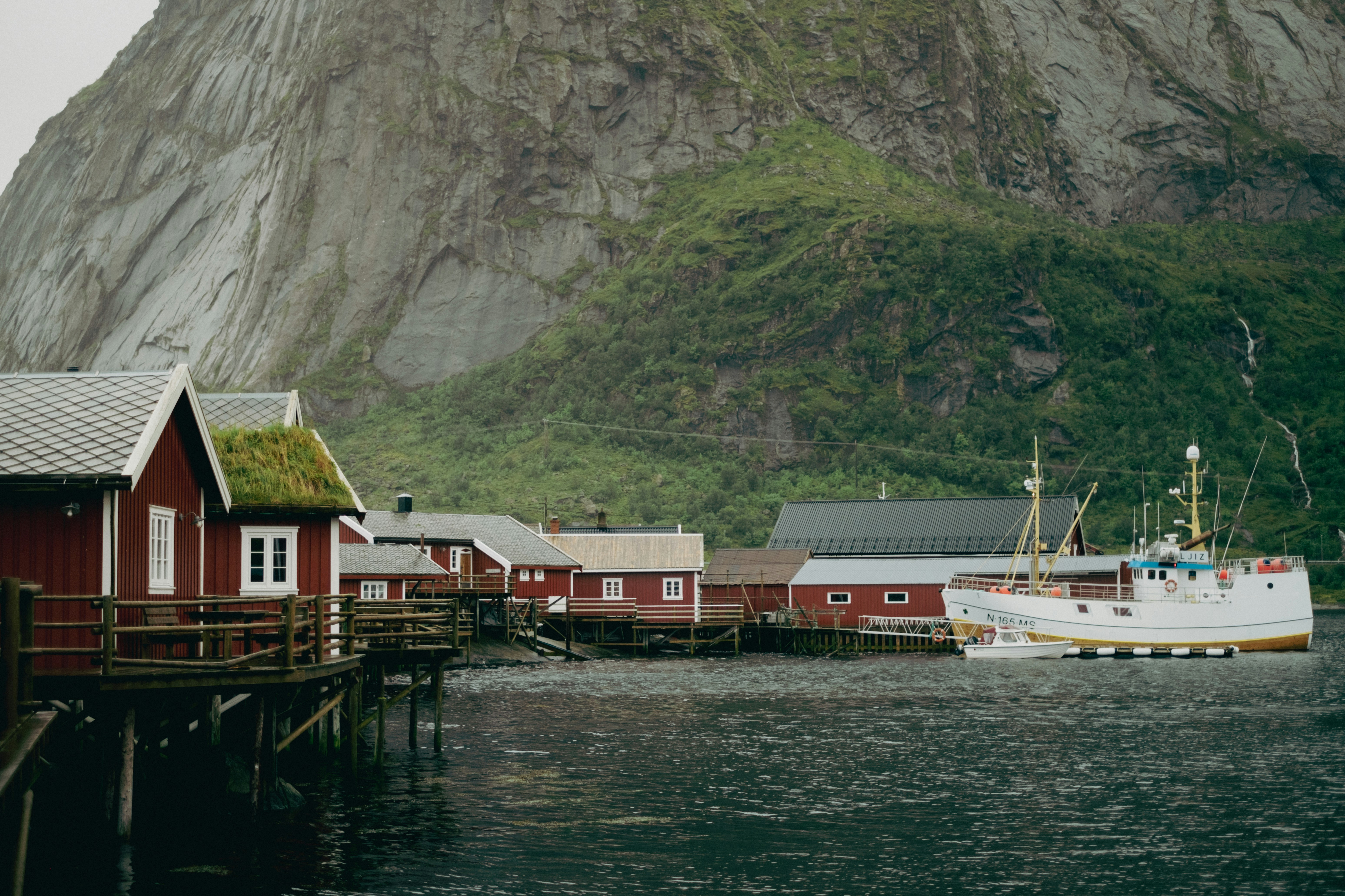 a boat docked at a dock in front of a mountain, Fishing village in Lofoten, Norway. Fishing boat and foggy weather with mountains in the background.
