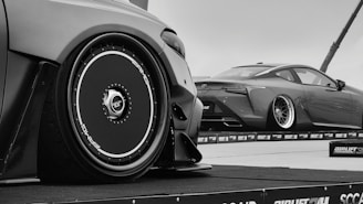 A black and white photograph featuring two sleek, customized sports cars with distinctive low-profile tires and glossy bodywork. The cars are parked on an elevated platform, likely part of a car show or exhibition. The focus is on the detailed design of the rims and the aerodynamic shape of the vehicles.