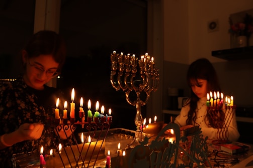 Two people are lighting candles on menorahs in a dimly lit room, creating a warm and intimate atmosphere. The menorahs are filled with colorful candles and are centrally placed, casting soft light around the room. The individuals appear to be engaged in a holiday tradition.