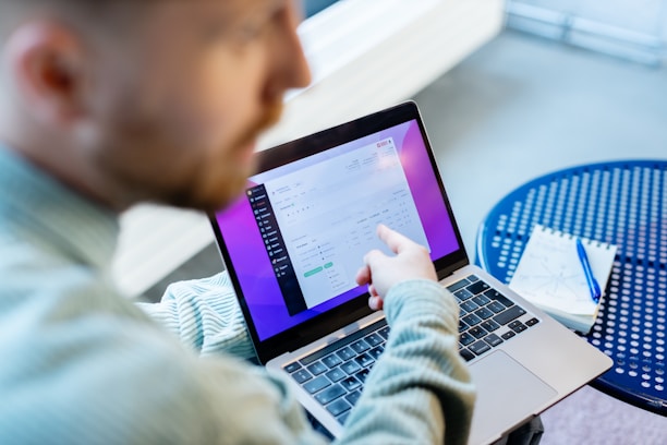 A person sitting at a round blue table, using a laptop. The laptop screen displays a digital interface with a purple sidebar and various data entries. The person is pointing at the screen, possibly explaining or highlighting something. A notepad and pen lie on the table, suggesting a work or study setting.