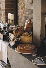 A rustic kitchen scene featuring various kitchen items on a counter next to a stone wall, including a freshly baked pie on a white ceramic tray, wooden utensils in a basket, candles, glassware, and ceramic jugs. The setting emphasizes a cozy and homely atmosphere with a vintage aesthetic.