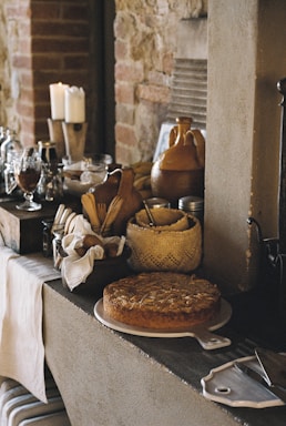 A rustic kitchen scene featuring various kitchen items on a counter next to a stone wall, including a freshly baked pie on a white ceramic tray, wooden utensils in a basket, candles, glassware, and ceramic jugs. The setting emphasizes a cozy and homely atmosphere with a vintage aesthetic.