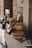 A rustic kitchen scene featuring various kitchen items on a counter next to a stone wall, including a freshly baked pie on a white ceramic tray, wooden utensils in a basket, candles, glassware, and ceramic jugs. The setting emphasizes a cozy and homely atmosphere with a vintage aesthetic.