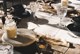 A rustic table setting with a mixture of natural and elegant elements. Glass goblets and dark ceramic bowls rest on a wooden table covered with a light linen runner. A jar of brown sugar sits alongside decorative straw and candles, creating a cozy and inviting atmosphere.