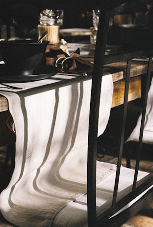 A rustic wooden dining table is set with a black bowl and a spoon on a matte black plate. A white table runner cascades over the table, creating subtle waves and shadows. A brown napkin, partially visible, adds contrast beside the bowl. In the background, glass jars and other tableware are slightly blurred, enhancing the focus on the foreground elements.