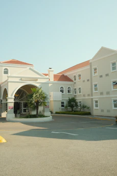 A beige hotel building with red-tiled roofs has an entrance featuring an arched design. The facade has several windows, and there are small palm trees and greenery around the entrance area. The ground is paved and there's a driveway leading to the entrance.