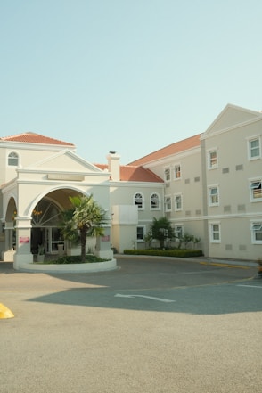 A beige hotel building with red-tiled roofs has an entrance featuring an arched design. The facade has several windows, and there are small palm trees and greenery around the entrance area. The ground is paved and there's a driveway leading to the entrance.