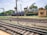 A railway scene featuring multiple train tracks intersecting and running parallel to each other. A green and yellow train marked 'Indian Railways' is positioned on one track, while a blue train labeled 'Indian Railways' is visible in the distance. The setting includes a small beige building to the right and some trees and industrial structures in the background, under a partly cloudy sky.