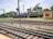 A railway scene featuring multiple train tracks intersecting and running parallel to each other. A green and yellow train marked 'Indian Railways' is positioned on one track, while a blue train labeled 'Indian Railways' is visible in the distance. The setting includes a small beige building to the right and some trees and industrial structures in the background, under a partly cloudy sky.
