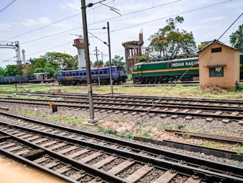 A railway scene featuring multiple train tracks intersecting and running parallel to each other. A green and yellow train marked 'Indian Railways' is positioned on one track, while a blue train labeled 'Indian Railways' is visible in the distance. The setting includes a small beige building to the right and some trees and industrial structures in the background, under a partly cloudy sky.