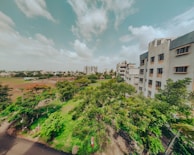 The image captures a view of an urban area with residential buildings on the right and lush green trees and grass in the foreground. The sky is partly cloudy, adding a serene, spacious feel. Farther in the background, more buildings and a vast open field are visible, contributing to a balanced mix of nature and urban development.
