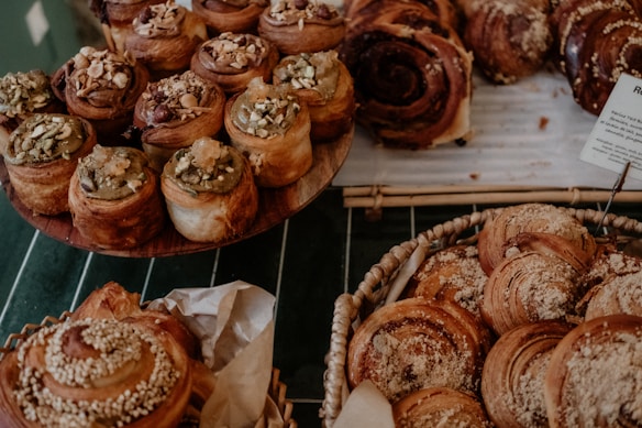 A collection of freshly baked pastries, including rolls and buns with various toppings like nuts and seeds, displayed on wooden and wicker trays. The image conveys a cozy and inviting atmosphere, emphasizing the textures and colors of the baked goods.