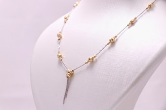 A delicate chain necklace displayed on a white mannequin bust. The necklace features gold bead elements distributed evenly along its length, with a central bead from which two chains hang down symmetrically.