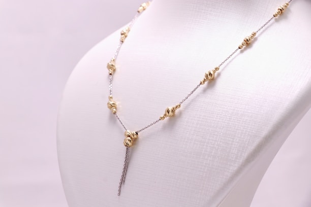 A delicate chain necklace displayed on a white mannequin bust. The necklace features gold bead elements distributed evenly along its length, with a central bead from which two chains hang down symmetrically.