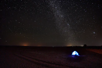 A vast, star-filled night sky stretches across the horizon, showcasing the Milky Way galaxy. A lone, illuminated tent stands in an open desert landscape, providing a striking contrast to the dark surroundings.