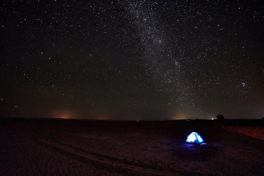 A vast, star-filled night sky stretches across the horizon, showcasing the Milky Way galaxy. A lone, illuminated tent stands in an open desert landscape, providing a striking contrast to the dark surroundings.