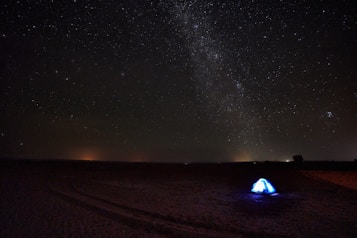 A vast, star-filled night sky stretches across the horizon, showcasing the Milky Way galaxy. A lone, illuminated tent stands in an open desert landscape, providing a striking contrast to the dark surroundings.