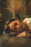 A person lies down with their head resting on a wooden table, surrounded by a potted plant with dried branches. Soft light and shadows create a tranquil atmosphere in the room.