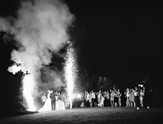A black and white photograph capturing a nighttime outdoor event with a large crowd gathered. Fireworks are ascending, illuminating the scene. In the foreground, a couple stands near a table, possibly the focus of the event.