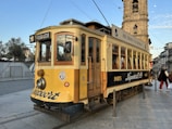 A vintage yellow and black tram is situated on a city street. The tram has a number '18' and the destination 'Passeio Alegre' displayed. It features wooden details and large windows, with passengers visible inside. In the background, there is a historic stone tower and people walking alongside. The sky is clear and blue, suggesting a calm day.