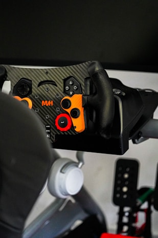 A close-up view of a gaming steering wheel with a carbon fiber texture. The wheel includes various buttons in colors such as orange and red, designed for game controls. It is mounted on a racing simulator setup, which also includes pedals visible in the background.