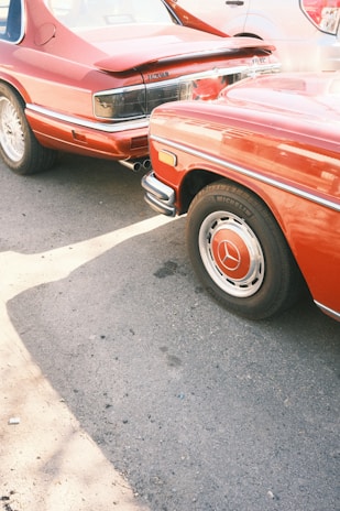 Two vintage cars parked closely together on a paved surface. The cars are both red, with one displaying the Jaguar emblem and the other featuring a Mercedes emblem. They are captured from the rear and side angles, highlighting their distinctive design details.