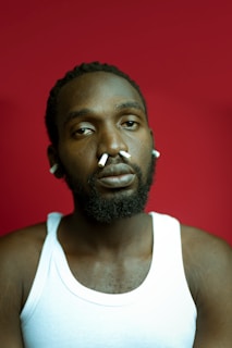 A person with a thoughtful expression wears a white tank top against a solid red background. Pieces of cotton or similar material are inserted in their ears and nostrils.