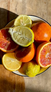 A bowl filled with various citrus fruits, including lemons, oranges, and blood oranges, placed on a wooden surface. The fruits are sliced to reveal their juicy and vibrant interiors, which catch the sunlight and create a warm, inviting atmosphere.