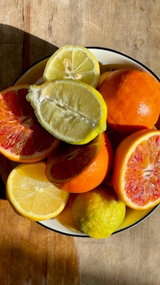 A bowl filled with various citrus fruits, including lemons, oranges, and blood oranges, placed on a wooden surface. The fruits are sliced to reveal their juicy and vibrant interiors, which catch the sunlight and create a warm, inviting atmosphere.