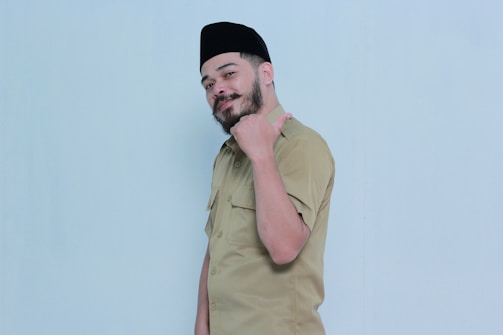 A man with a beard wearing a khaki uniform and a black cap poses with a confident expression. He gestures with his thumb pointing towards himself against a plain light blue background.