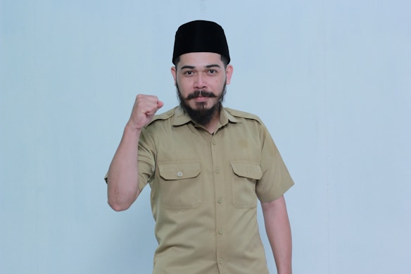 A man wearing a light brown uniform shirt and a black cap is posing against a plain background. He has a beard and mustache, and his right fist is raised in a gesture of determination or solidarity.