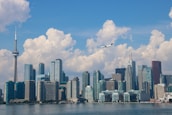 A city skyline features a range of modern skyscrapers along a waterfront, dominated by a very tall observation tower. A plane flies in the blue sky, which is filled with large, fluffy clouds.