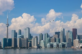 A city skyline features a range of modern skyscrapers along a waterfront, dominated by a very tall observation tower. A plane flies in the blue sky, which is filled with large, fluffy clouds.