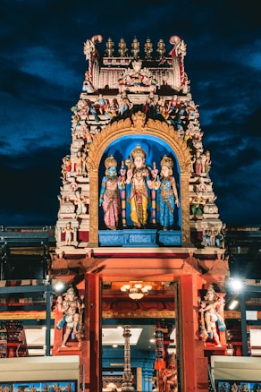 An intricately designed Hindu temple facade featuring vibrant statues of deities adorned with vivid colors and elaborate detailing, set against a dramatic evening sky.