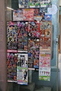 A window display covered with colorful posters featuring a variety of collectible figurines and merchandise. The posters depict animated characters and promotional images for toys, with text in different languages including Japanese. The items appear to be popular franchises, showcasing a vibrant and lively array of designs.