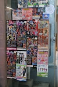 A window display covered with colorful posters featuring a variety of collectible figurines and merchandise. The posters depict animated characters and promotional images for toys, with text in different languages including Japanese. The items appear to be popular franchises, showcasing a vibrant and lively array of designs.