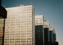 A series of tall, modern office buildings with reflective glass facades and sharp, rectangular structures under a clear blue sky.